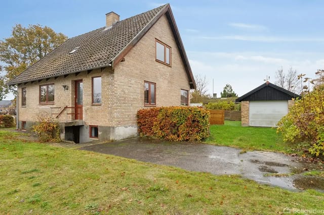 A yellow brick house with a red roof stands next to a garage, surrounded by a green garden with bushes and trees in autumn foliage.