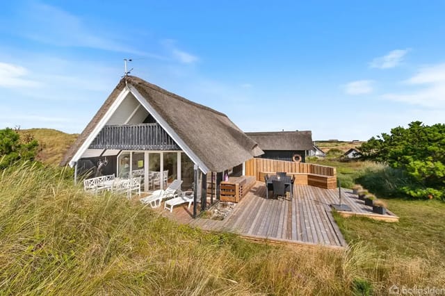 Thatched house in dune landscape with large wooden terrace, grill, and garden furniture.