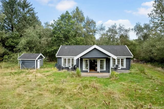 A gray house with white windows stands quietly on a lawn surrounded by trees. To the left is a small shed. The sky is blue with scattered clouds.