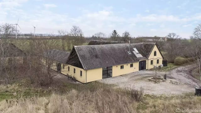 Yellow farmhouse with gray roof in a rural area surrounded by trees and fields. A gravel courtyard is seen in front of the house. Wind turbines are visible in the distance.