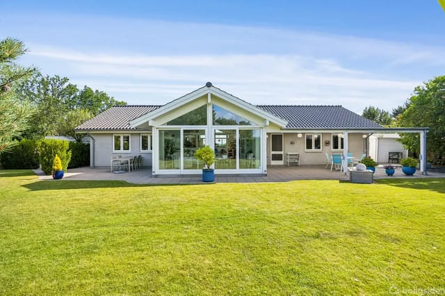 A modern house with large windows and a terrace in a well-kept garden with green trees and bushes. Blue pots with plants on the terrace.