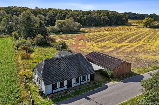 A house with a black roof and white walls stands next to a barn on a country property surrounded by fields and a forest in the background, in sunny weather.