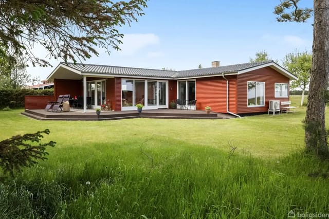 A red wooden house stands on a green lawn with large windows and a wooden terrace with furniture, surrounded by trees and bushes in a rural area.