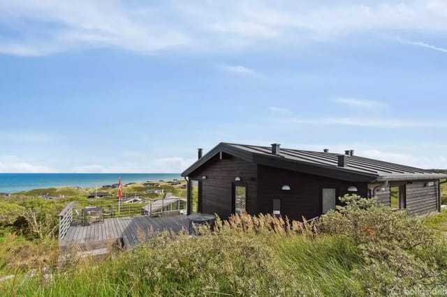 A black wooden house stands quietly in a lush dune landscape with a view of the sea. In the foreground, there is grass and bushes, and the sea is glimpsed in the background.