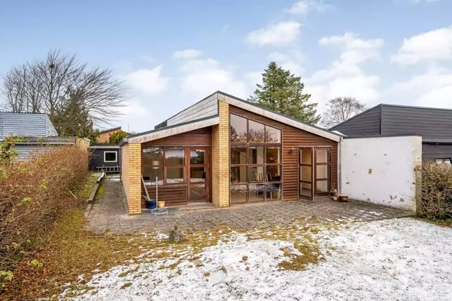 A modern house with large windows and a sloping roof stands in a snow-covered garden. Around the house are bushes and trees under a blue sky with clouds.