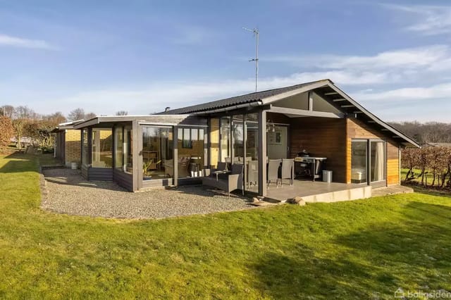 A modern holiday home stands quietly on a well-kept lawn in a sunny landscape. The house features large windows and a small terrace, surrounded by bushes and open fields.