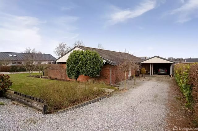 A red brick house with a brown roof, surrounded by a garden, stands next to a gravel driveway. At the end of the driveway is a carport.