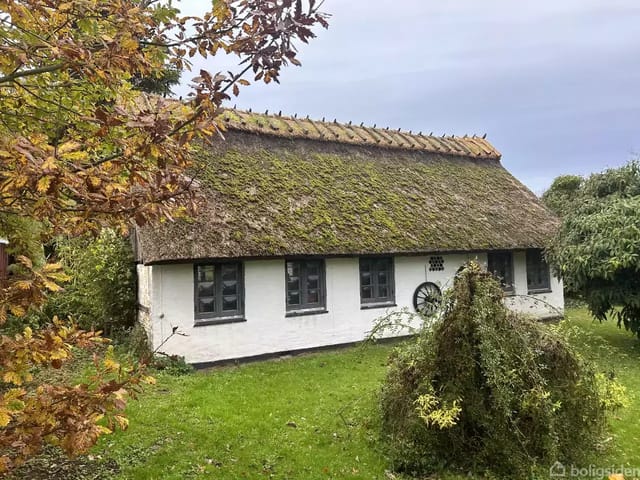 A thatched house with moss on the roof stands quietly in a lush garden surrounded by trees and lawn on a gray cloudy day.