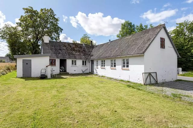 A white farmhouse with a black roof stands in a green garden with a lawn. The house has several small windows and is under a blue sky with scattered clouds.