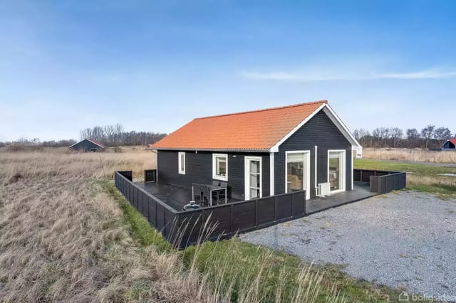 A black wooden house with a red roof surrounded by wild grass and open landscape. It has a fenced terrace and a gravel area in front suggesting a courtyard.