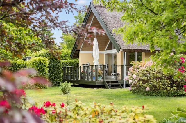 A thatched house with a wooden terrace surrounded by a lush garden with flowering bushes and trees. On the terrace are furniture and a folded parasol, creating a peaceful atmosphere.