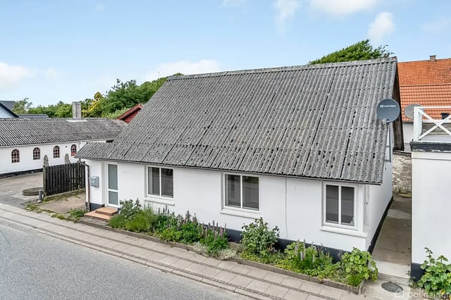 A white detached house stands on a quiet street with a sloped roof, satellite dish, small shrubs, and a paved walkway.