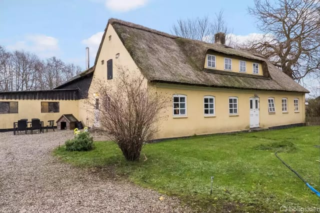 A yellow half-timbered house with a thatched roof stands in a courtyard surrounded by gravel and grass. Next to the house is an outdoor area with garden furniture and a small shed.