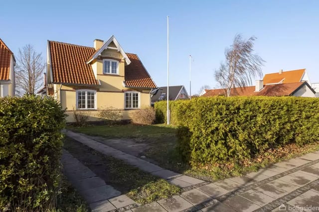 A yellow house with a red roof located in a garden with a paved driveway; surrounded by a green hedge. Clear blue sky in the background.