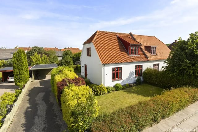 A white house with a red tile roof stands in a well-kept garden with green bushes. A driveway leads to a carport next to the house in a residential neighborhood.