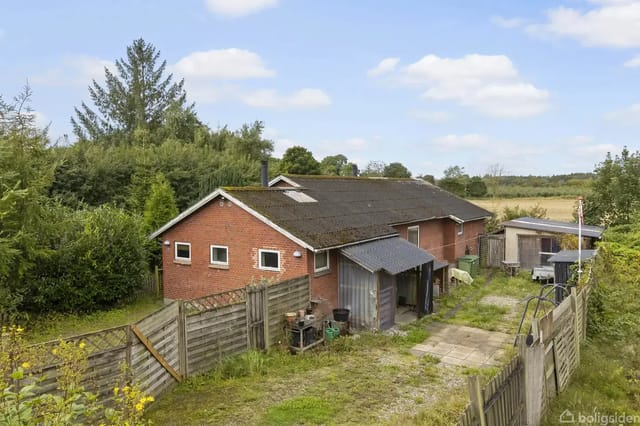 A red brick building with a black roof stands in a garden enclosed by a wooden gate fence. Trees and bushes surround the building in a rural area with open fields in the background.