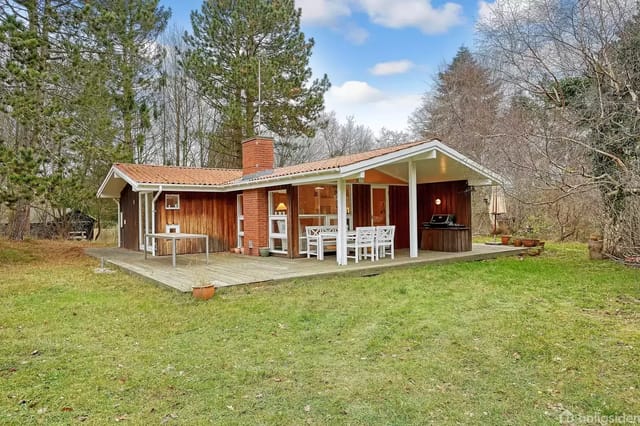 A wooden house with a red roof on a lawn, surrounded by forested areas with tall trees.