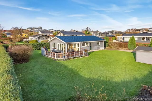 A house with a wooden terrace stands on a green lawn, surrounded by a garden in a residential area with several houses in the background under a blue sky.