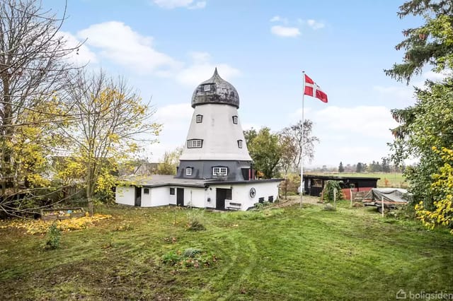 A white, old-fashioned mill tower stands on a lawn surrounded by trees, with a Danish flag waving from a flagpole next to it.