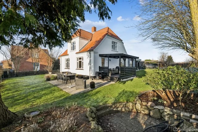 A white house with a red roof stands in a lush garden. There is a terrace with garden furniture, surrounded by green lawn and bushes, under a blue sky.