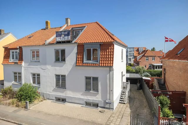 A two-story building with a red tile roof for sale in a residential area. A ladder is on the side, and a Danish flag is raised nearby.