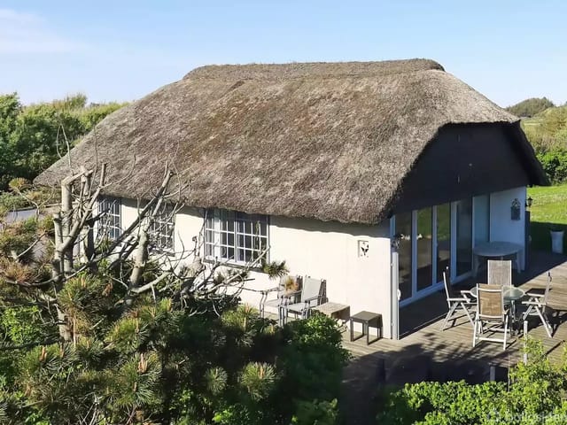 A thatched-roof summer house stands quietly in sunlight surrounded by green bushes and trees. In front of the house is a wooden terrace with garden furniture, creating an inviting outdoor atmosphere.