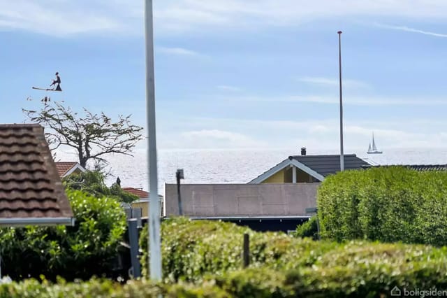 Man jumping from a tree over rooftops with sea and sailboat in the background. Green hedges frame the foreground under a blue sky.