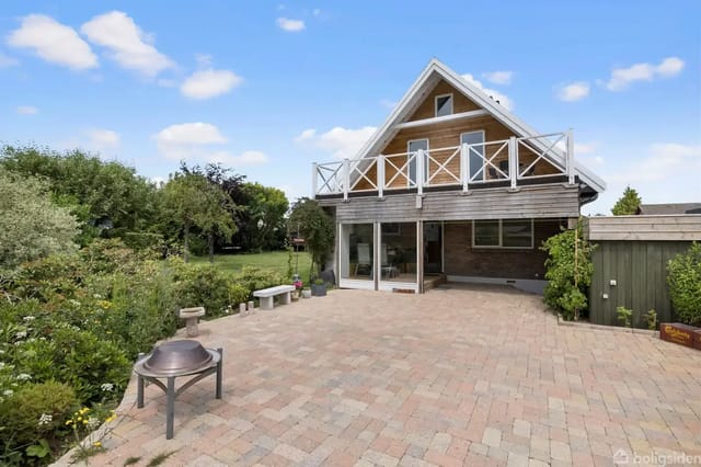 A wooden house with a balcony and windows, standing on a paved terrace. Surrounded by a lush garden with bushes and trees under a bright blue sky.