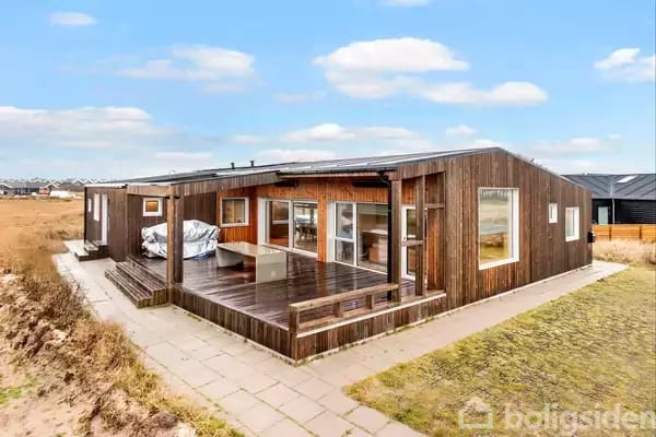 A modern wooden summer house located on a paved area by a beach. The house has large windows and a spacious terrace with a canopy, surrounded by grass and sand dunes in the background.