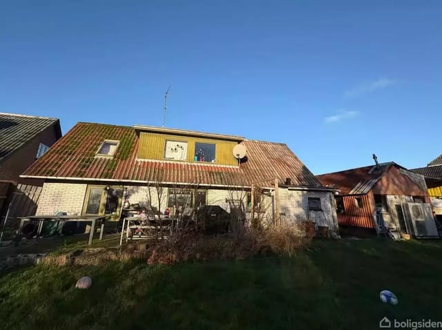 An old house with a sloping roof and white bricks stands in a garden with bushes and grass. Two windows are placed at the top, and a satellite dish is mounted on the roof.