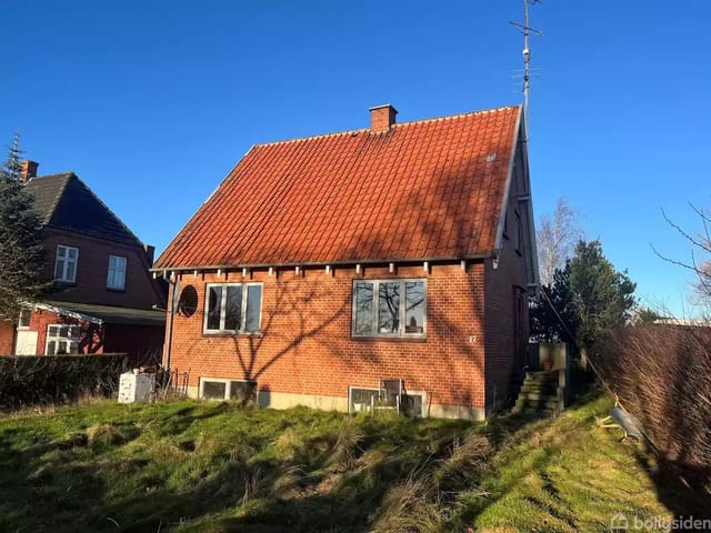 Red brick house with sloped roof in a green garden area. The sun is shining, and tree shadows hit the house facade. Blue sky forms the background.