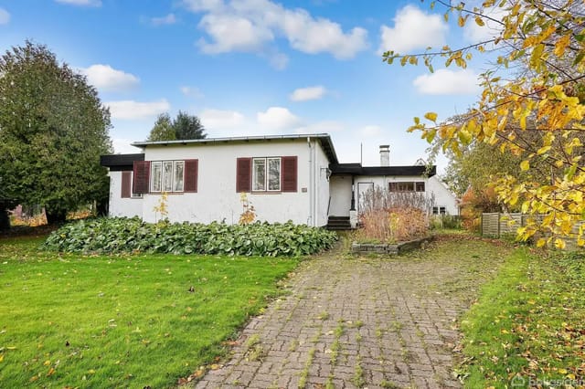A white house with brown shutters stands in a garden with green lawn and trees. A paved path leads to the entrance under a blue sky.