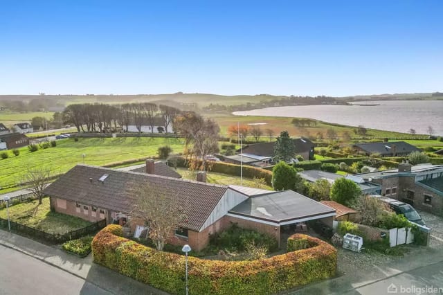 House with garden surrounded by hedge, located by a road. In the background, a lake and open green areas under a blue sky.