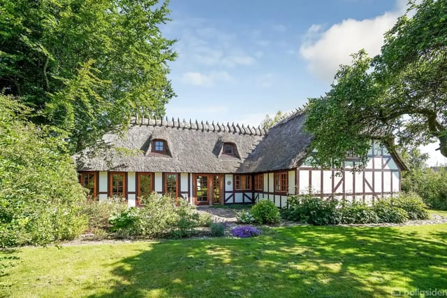 Thatched half-timbered house surrounded by a green garden with flower beds and bushes. Detailed wood structures highlighted by sunlight. The forest encircles the property, creating a natural backdrop.