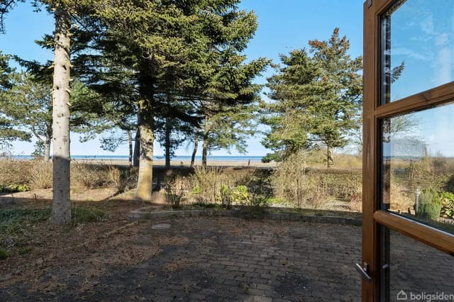 A door opens onto a cobblestone terrace. In the background, a garden with tall trees and a glimpse of the sea under a blue sky.