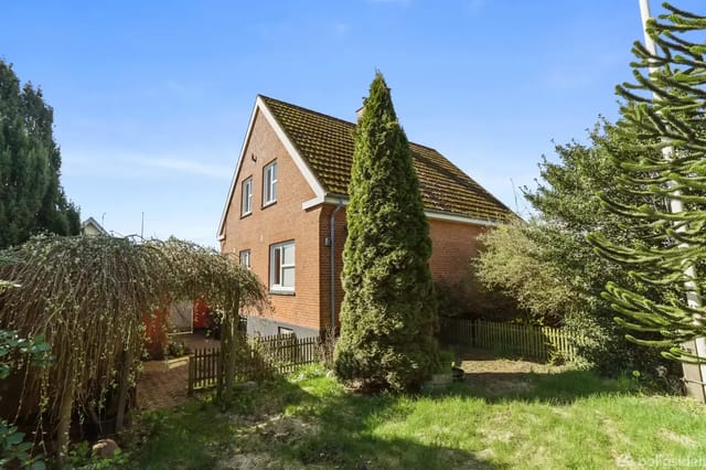 A red brick house stands quietly surrounded by a green garden with dense bushes and trees under a clear blue sky.