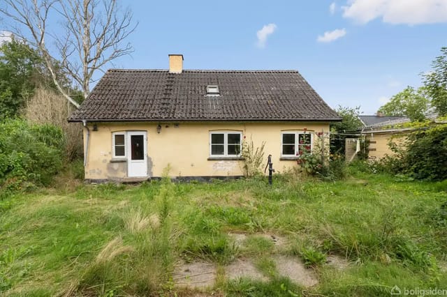 A yellow-painted house stands quietly on an overgrown lawn, with a black roof, several windows, and a door to the left surrounded by bushes and trees.