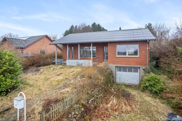 A red brick house on a slope with a garage in the basement. Surrounding the house are bushes and grass, with more trees and another house in the background.