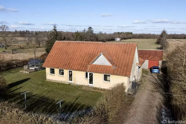 A house with a red roof in a rural setting surrounded by fields and trees. There is a gravel road beside the house and a car in the driveway.