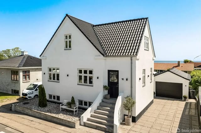 White house with black tiled roof on a paved lot, number '16' by the door, garden with plants and a small garden house in the background.