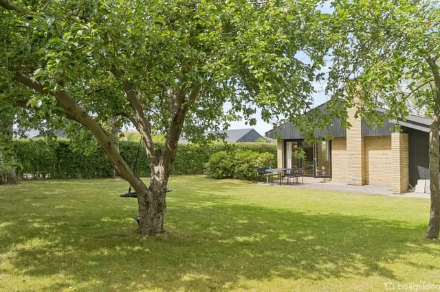 A bright brick house with number 13 stands quietly among green trees and bushes, flanking a paved driveway leading to a carport.