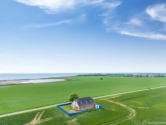 A solitary brick house stands quietly surrounded by lush green fields under a clear blue sky by the coast. A road runs close by the building.