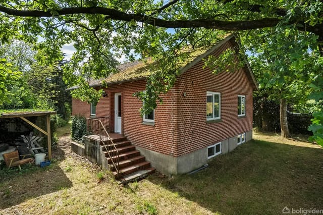 Red brick house surrounded by green trees and a lawn. A staircase leads to the main entrance, with a shed and firewood to the left.