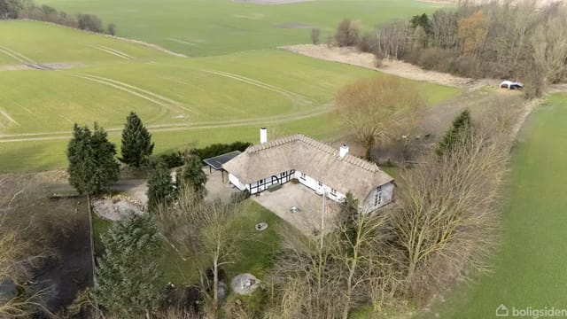 Thatched half-timbered house in an open, rural area surrounded by fields and trees.