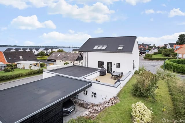 House with white facade and dark roof, terrace with grill and furniture, located in a peaceful residential area near the coast, surrounded by green gardens and neighbors.