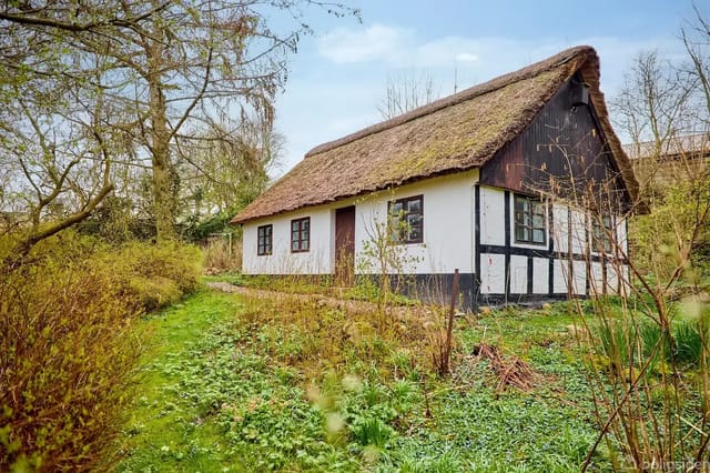 Half-timbered house with thatched roof in a lush garden surrounded by trees and bushes under a blue sky.