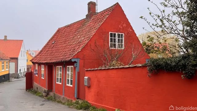 Red house with sloped roof on a cobblestone street, surrounded by bushes and colorful houses.