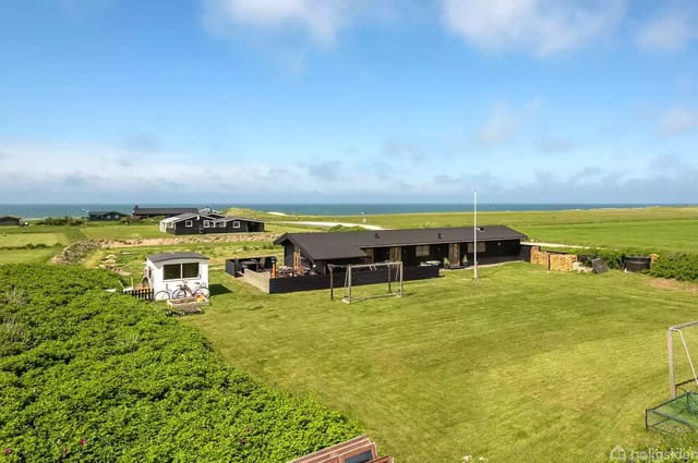 A black wooden summer house stands quietly in a green garden with a swing set, a few meters from the sea on a clear day. A small white caravan is seen to the left.