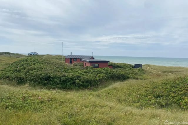 A red house stands quietly among green dunes by the sea. Clouds cover the sky, and there are no people present in the scenic landscape.