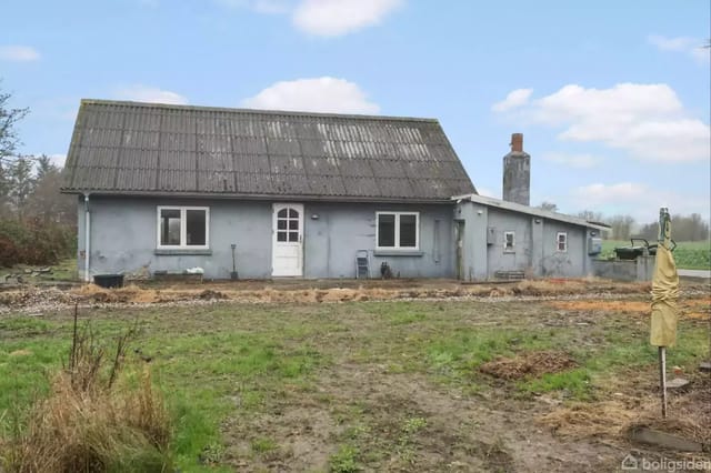 A gray house with a sloped roof stands quietly on a lawn. The house has a chimney and is surrounded by a prominent garden. The sky is blue with white clouds.